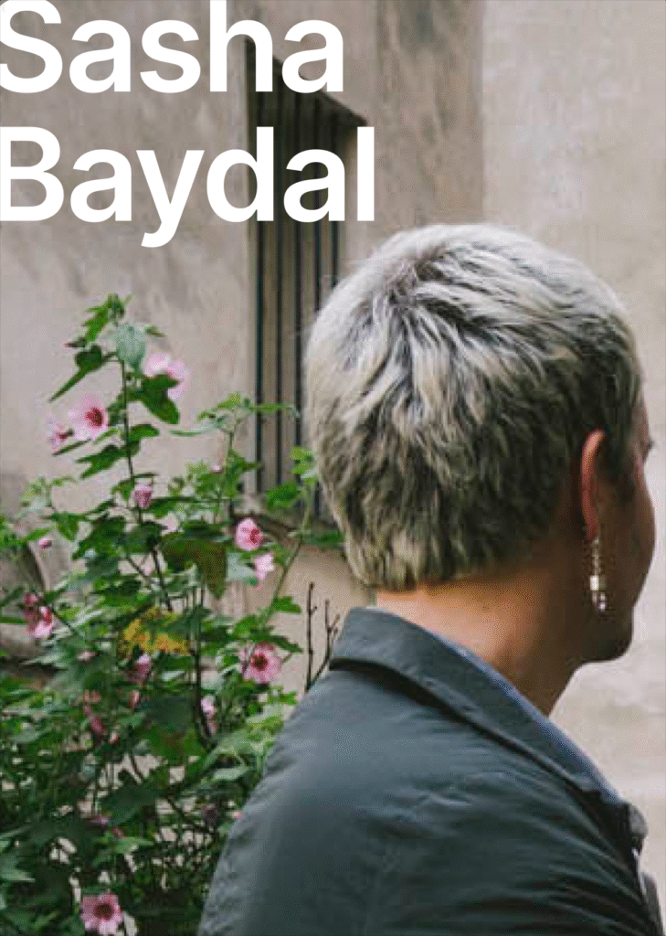 There is the picture of Sasha's head from the back. They are wearing an earring on the right ear. On the background there is a begue wall and green bushes. The name of the speaker is written in white letters "Sasha Baydal".