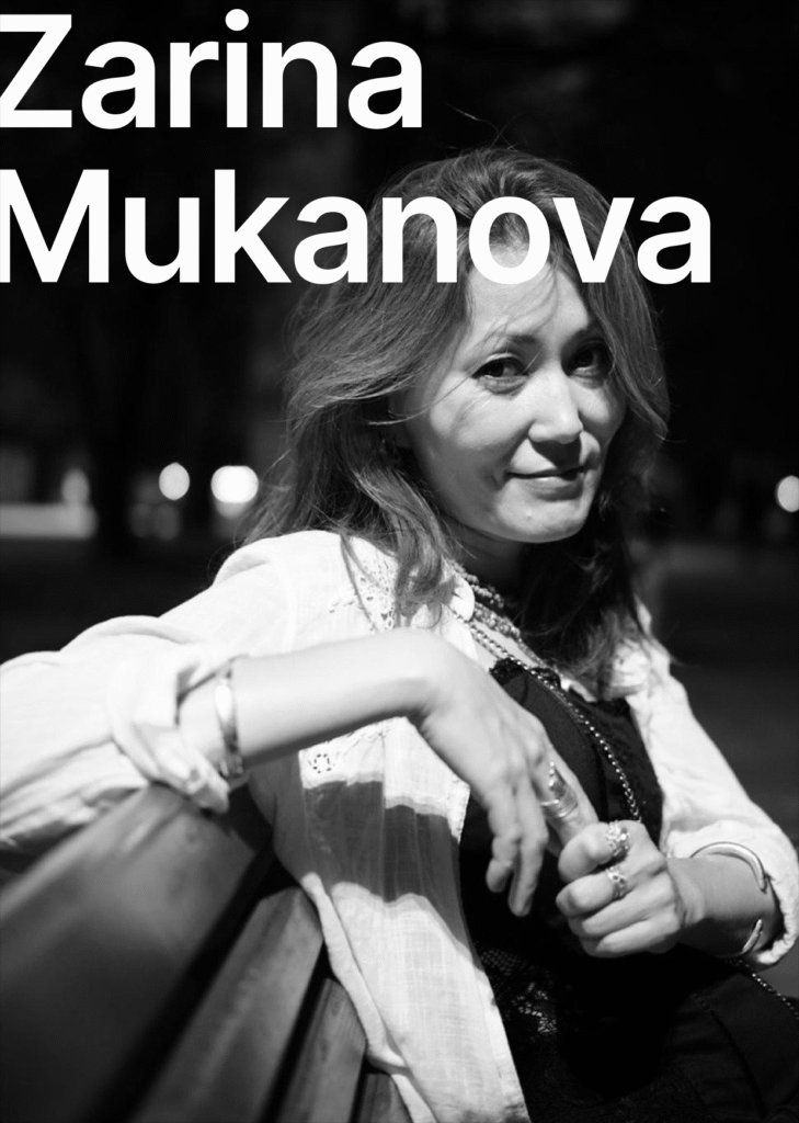 There is a black and white picture. The text says Zarina Mukanova. The person is sitting on the bench. She is wearing a white shirt and a black t-shirt. She has shoulder length hair. She has rings on her fingers and a longer necklace. She is smiling at the camera.