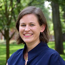 There is a colourful bright picture in the background. There is a person standing in front of the building. The person is wearing a blue dress and wears short brown hair. The person is smiling friendly.