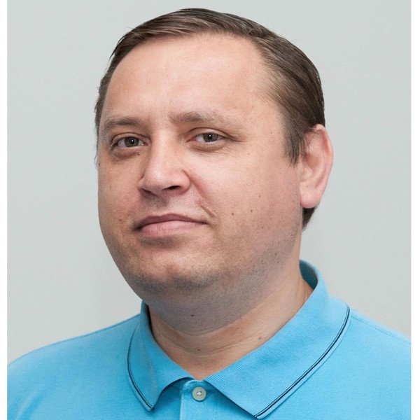 An image of a male person on a grey background. He is smiling friendly and has a short haircut and blue t-shirt on.