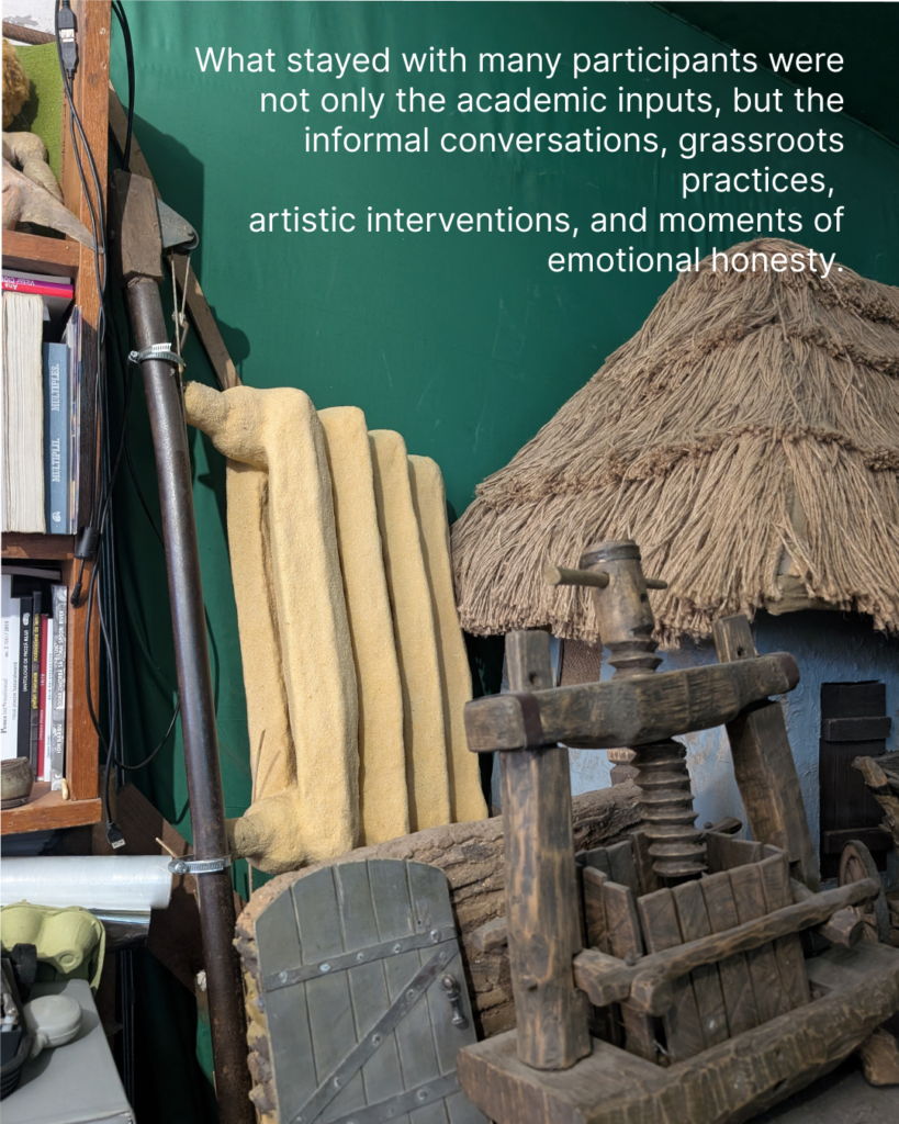 In the picture there are wooden hand-made objects. There is a small well, the entrance to the village house with the small fence, a house with a straw roof. The wall in the background is bright green, There is a text in teh background: what stayed with many participants were not only teh academic inputs, but the informal conversations, grassroots practices, artistic interventions, and moments of emotional honesty.