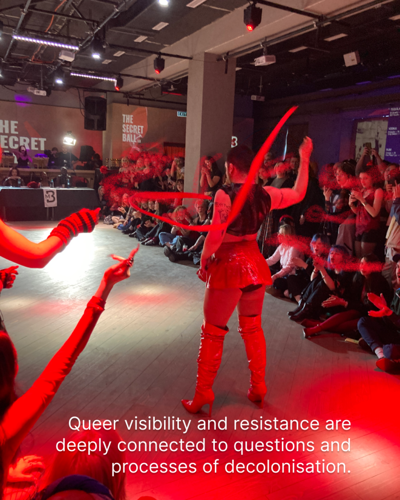 In the picture there is a performance, where the person is dancing. The light is red. There are a lot of people in the background, looking at the dancing person and cheering. The person is wearing knee high high heels, a skirt and a jacket. There is a text in the background: queer visibility and resistance are deeply connected to questions and processes of decolonisation.