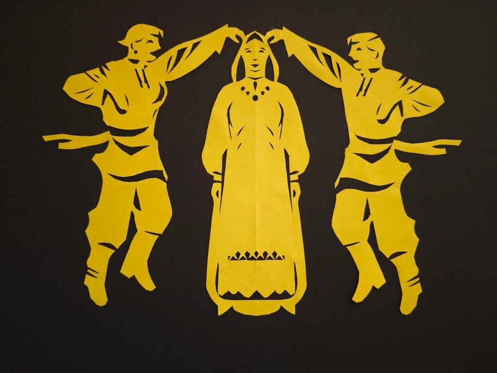 Der Scherenschnitt zum Buchstaben B für Bisexuell ist in der Farbe gelb. Es zeigt drei Personen. Die Personen rechts und links im Bild haben die Arme erhoben und scheinen um die mitllere Person herumzutanzen. Die beiden haben eine Oberteil an, um das an der Taille ein wehender Schal gebunden ist. Sie haben eine weite Hose und Stiefel an. Die Person links hat außerdem einen Ohrring und längere Wimpern. Die Person in der Mitte lächelt und trägt ein weites Kleid mit dekorativen Ornamenten, eine Kette und ein Tuch auf dem Kopf.