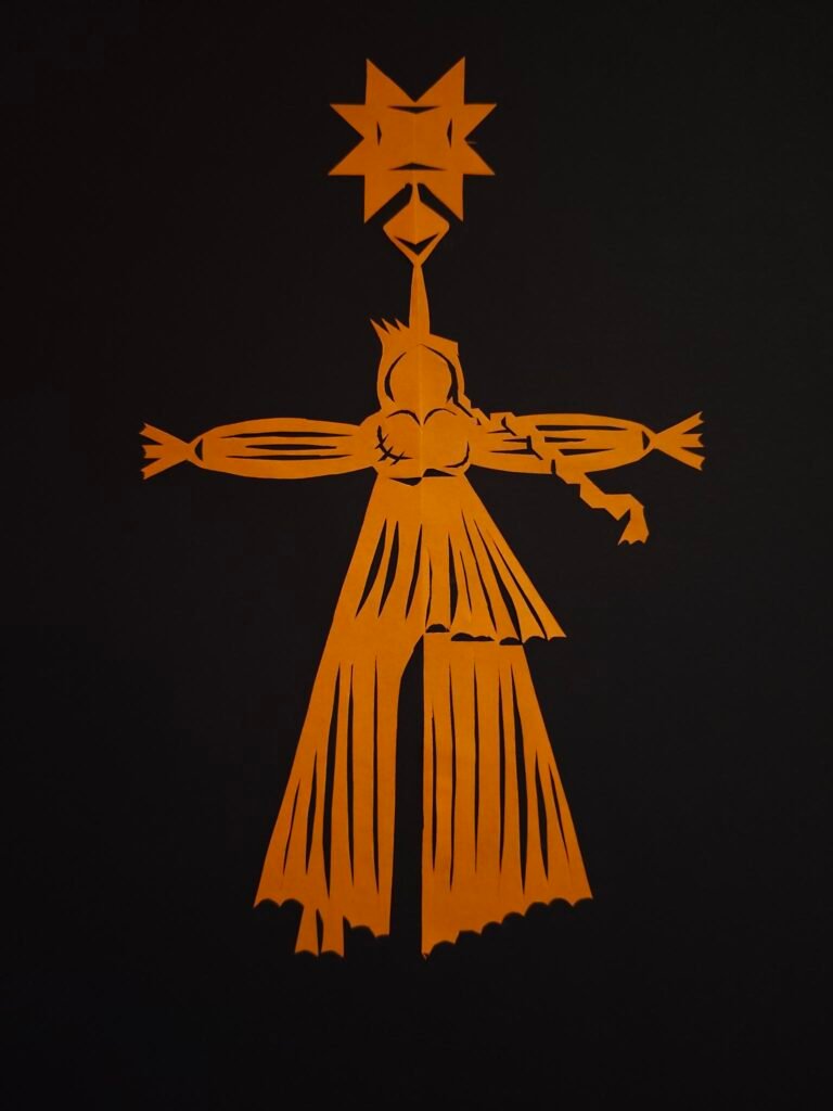 The paper cut-out representing the letter G for ‘gay’ is orange in colour. It depicts a person with outstretched arms and one leg extended, as well as an octagonal star on their head. The figure is wearing a flowing, pleated garment consisting of a top and loose-fitting trousers. The top has an accentuated chest area; two slits on the left-hand side could also be interpreted as scars. The figure has a sort of short fringe swept up on the left side and a long plait on the right.
