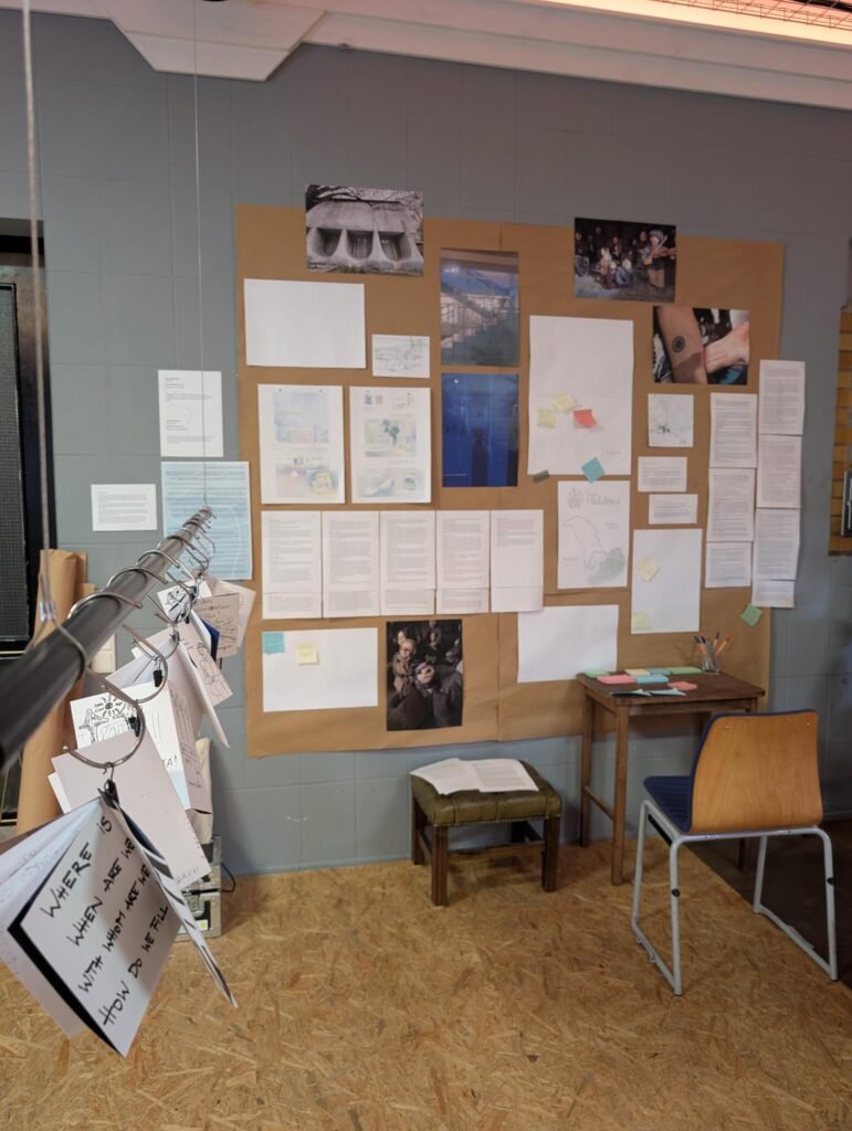 In the background a wall with pictures, texts and comics. next to the wall a table with colored small papers and a chair. In the front a pole with paper zines haning next to each other.