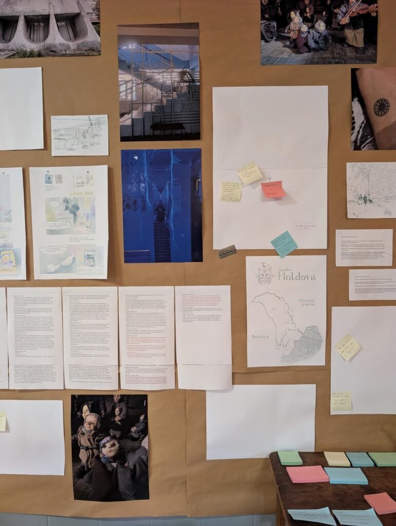 Description: In the background a wall with pictures, texts and comics. next to the wall a table with colored small papers and a chair. In the front a pole with paper zines haning next to each other.