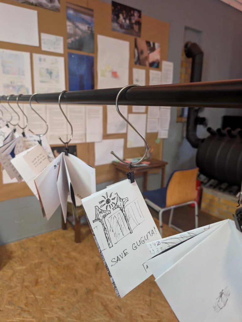 Description: In the background a wall with pictures, texts and comics. next to the wall a table with colored small papers and a chair. In the front a pole with paper zines haning next to each other.